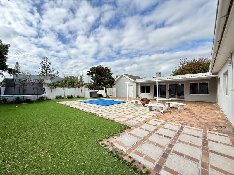 Spacious rear garden featuring a paved patio area, covered entertainment space, swimming pool with cover, and low-maintenance artificial lawn.
