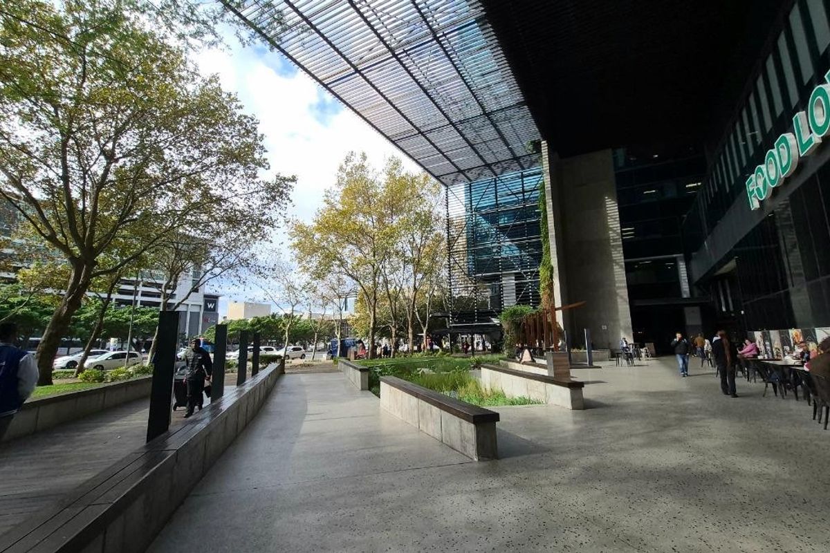 Modern commercial outdoor plaza featuring concrete paving, built-in seating, and a large metal louvered canopy adjacent to a glass-fronted building.
