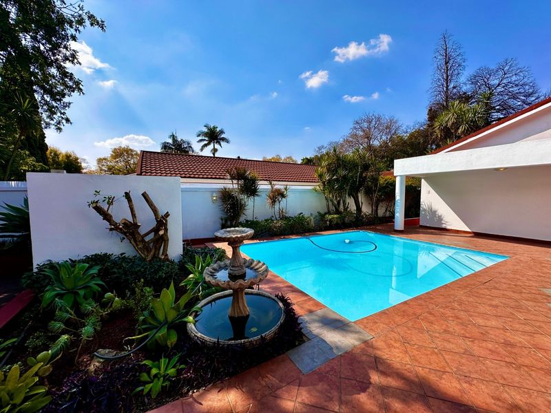 A well-maintained outdoor pool area featuring a tiled patio, a decorative stone fountain, and a covered patio section, set within a private garden.
