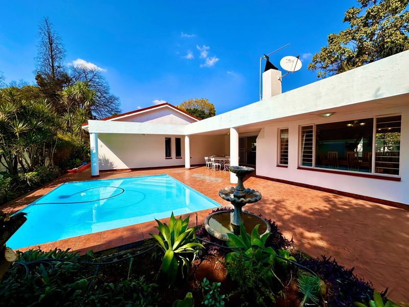 A well-maintained outdoor pool area featuring a tiled patio, a central stone fountain, and a covered entertainment space.