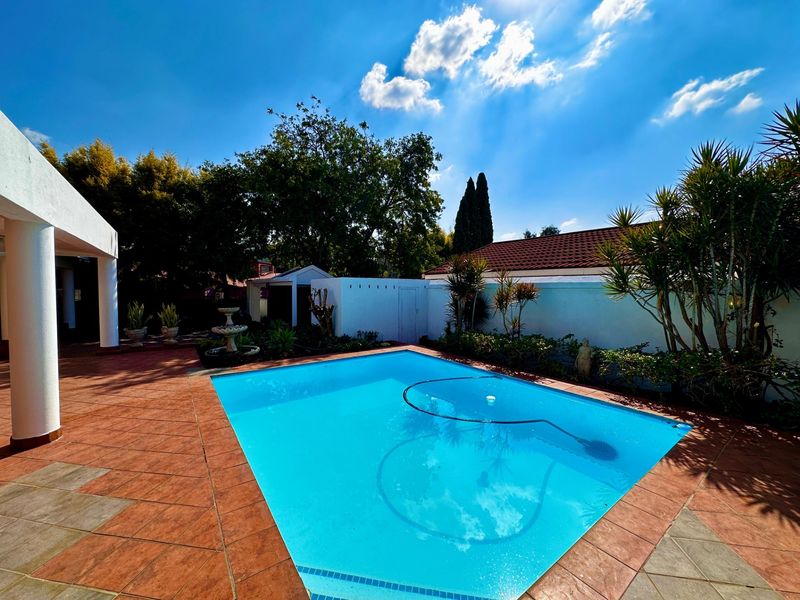 A well-maintained outdoor pool area featuring a tiled patio, surrounding boundary walls, and a covered patio section.