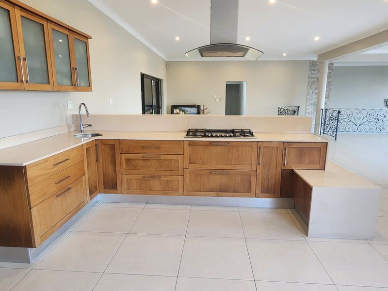 A spacious kitchen featuring wooden cabinetry, stone countertops, a gas hob with extractor fan, and tiled flooring, opening into a large living area.
