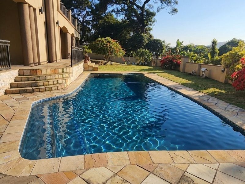 A well-maintained swimming pool area featuring stone paving, a retaining wall, and a metal balustrade, set within a landscaped garden.
