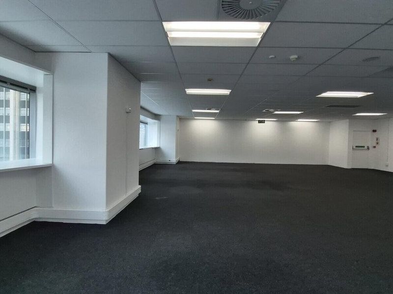 A spacious, open-plan commercial office area featuring a suspended ceiling, recessed lighting, and large windows providing natural light.