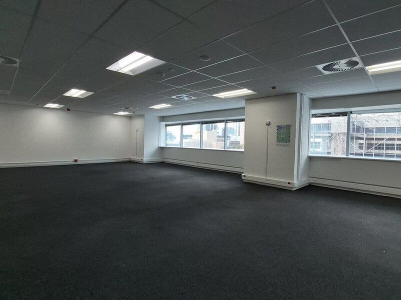 A spacious, open-plan commercial office area featuring a suspended ceiling, recessed lighting, large windows providing natural light, and dark carpeted flooring.