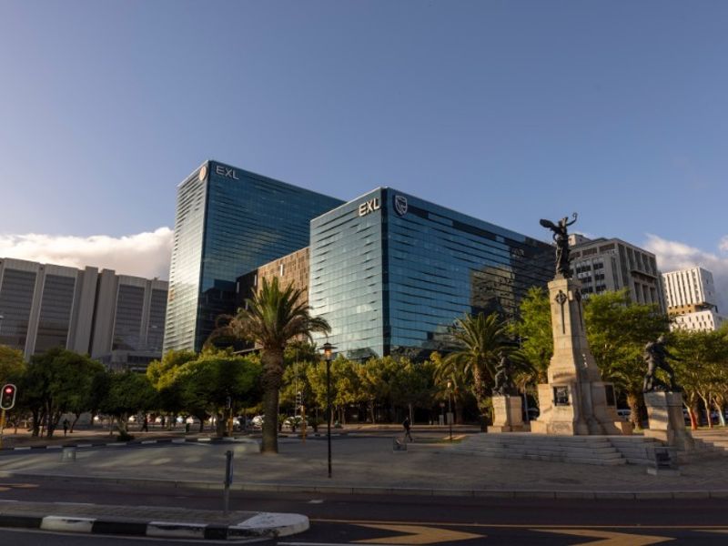 A professional exterior view of a modern glass-facade commercial office building situated near a public plaza and monument.