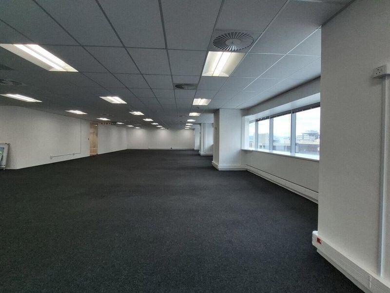 A spacious, open-plan commercial office area featuring suspended ceilings, recessed lighting, large windows for natural light, and dark carpeting.