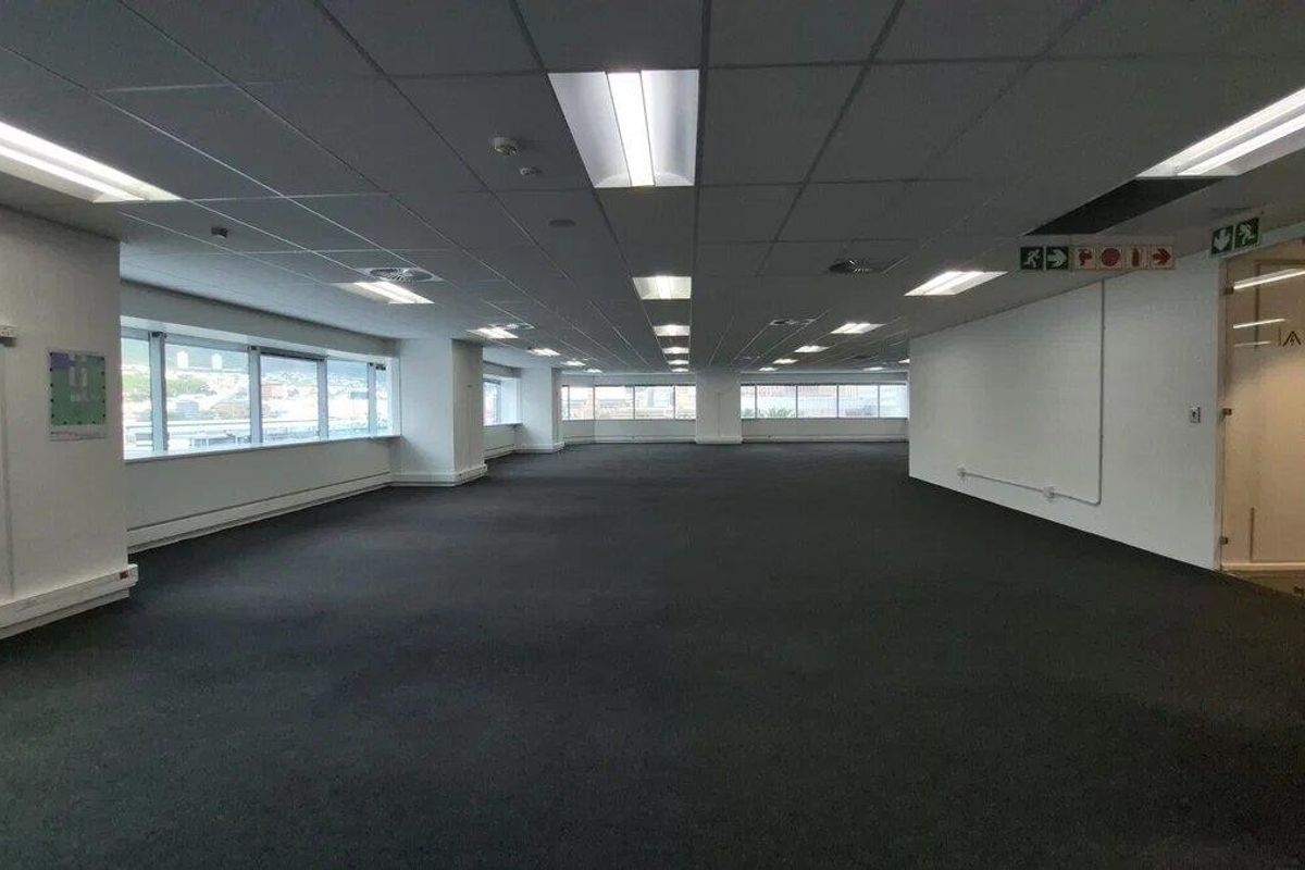 A spacious, open-plan commercial office area featuring suspended ceilings, recessed lighting, carpeted floors, and large windows providing ample natural light.