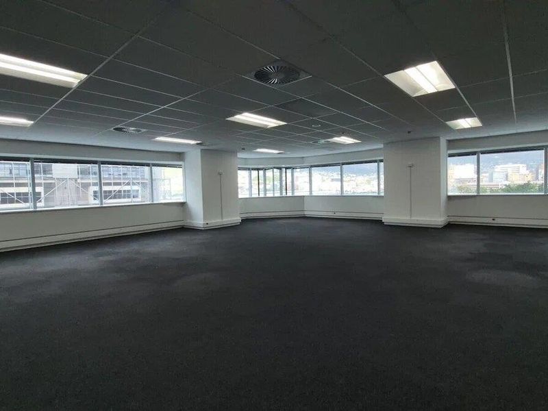 A spacious, empty open-plan office area featuring large windows, a suspended ceiling with recessed lighting, and carpeted flooring.