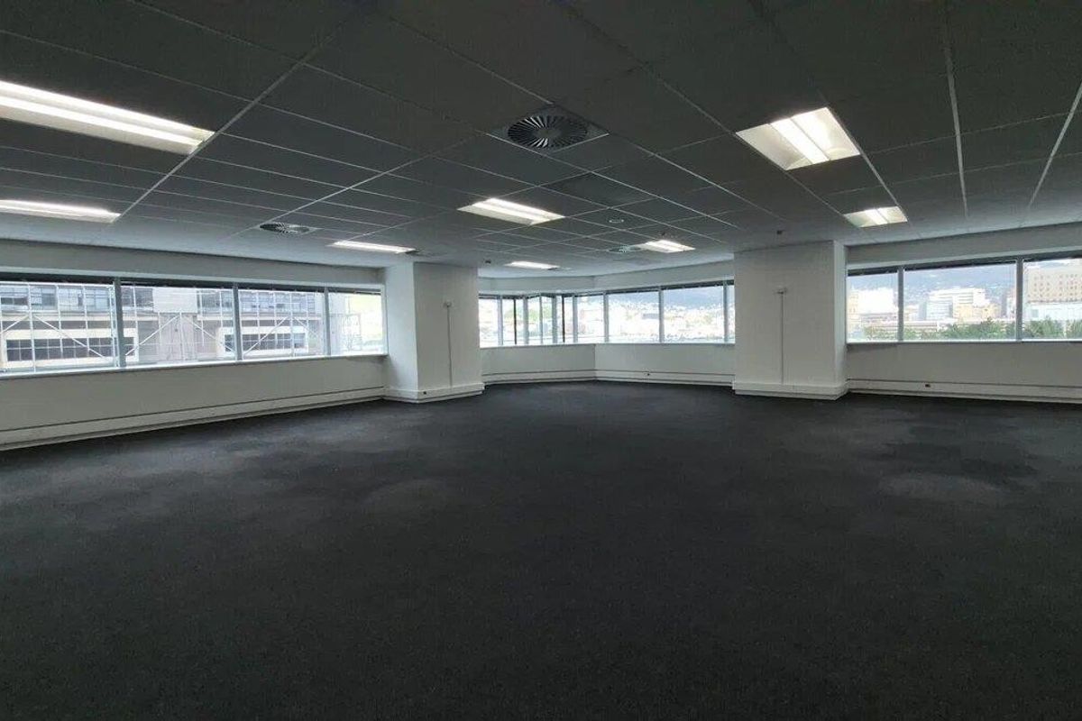 A spacious, empty open-plan office area featuring large windows, a suspended ceiling with recessed lighting, and carpeted flooring.
