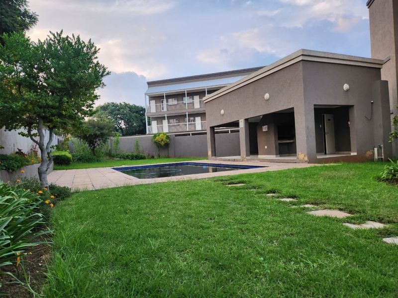 Communal pool area featuring a paved deck, covered patio with built-in braai, and surrounding garden space within an apartment complex.