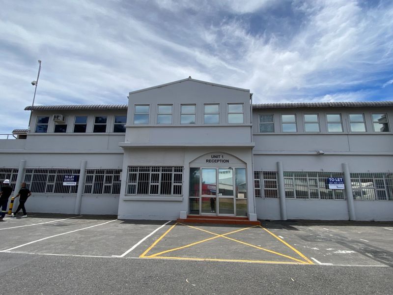 Front elevation of an industrial unit featuring a dedicated reception entrance, security-barred windows, and an asphalt-paved parking area.