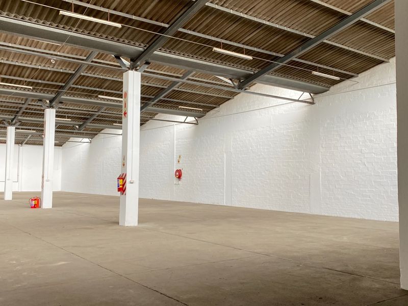 Spacious industrial warehouse unit featuring high ceilings with exposed steel trusses, durable concrete flooring, painted brick walls, and a large roller shutter door.