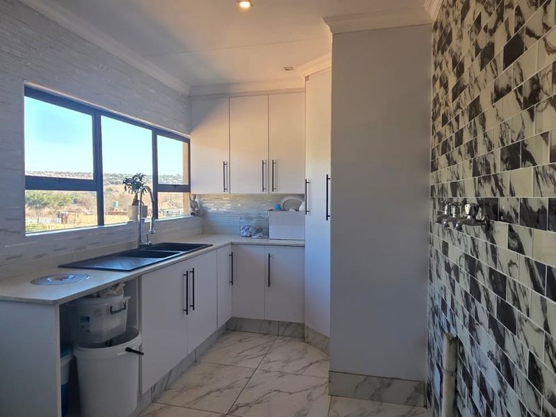 A clean and functional laundry room featuring modern white cabinetry, a double sink, and decorative tiled wall finishes with plumbing connections.