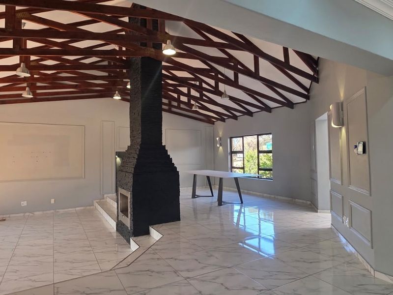 Spacious living area featuring high exposed timber roof trusses, a central stone-clad fireplace, and modern tiled flooring with elegant wall paneling details.