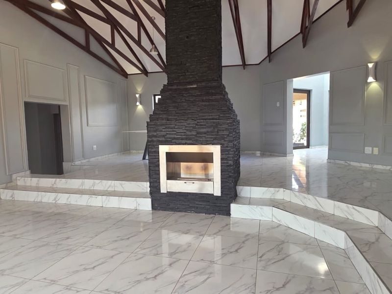 Spacious living area featuring a striking central stone fireplace, vaulted ceilings with exposed timber trusses, and elegant marble-look tiled flooring with raised levels.