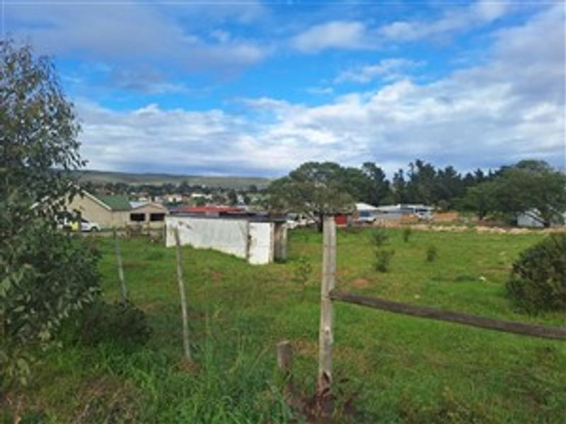 A vacant residential plot featuring a wire fence, wooden posts, and a small, weathered outbuilding structure set against a suburban landscape.
