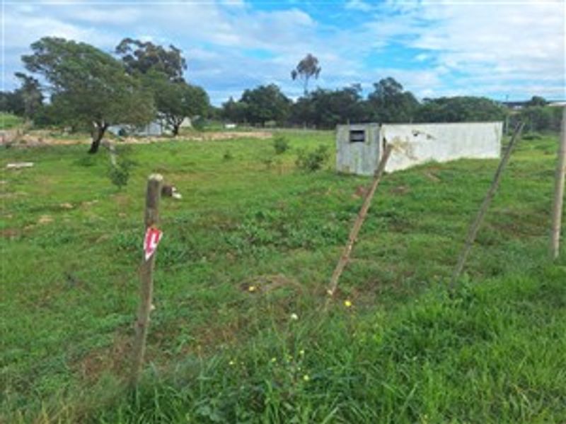 A view of a vacant residential plot featuring grassy terrain, perimeter wire fencing, and a small, weathered concrete outbuilding.