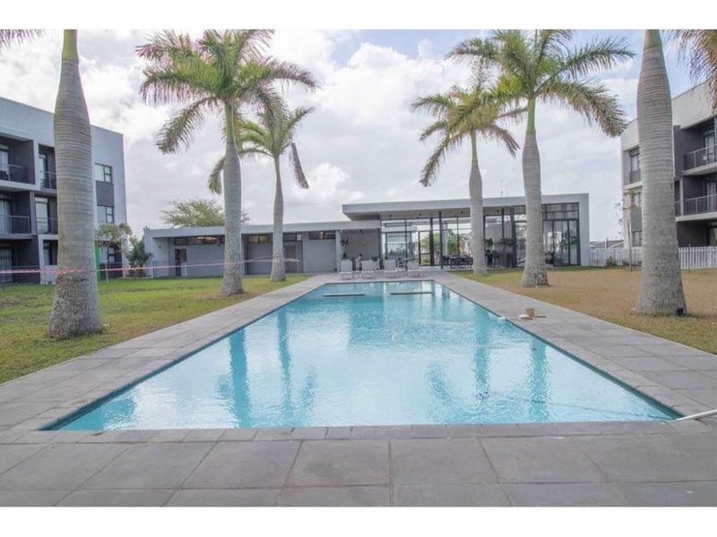 A well-maintained swimming pool area featuring a paved deck, surrounding garden with palm trees, and an adjacent modern clubhouse building.