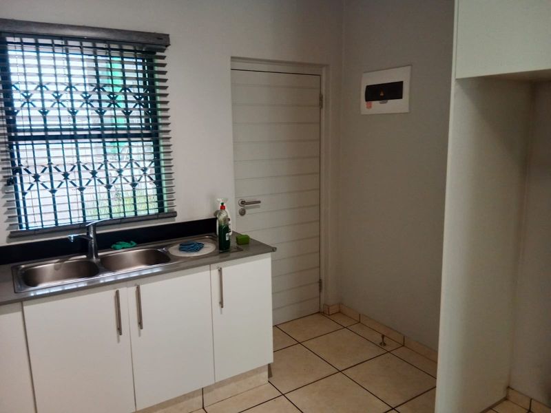 A functional scullery area featuring a double stainless steel sink, built-in white cabinetry, tiled flooring, and a secure window with burglar bars.