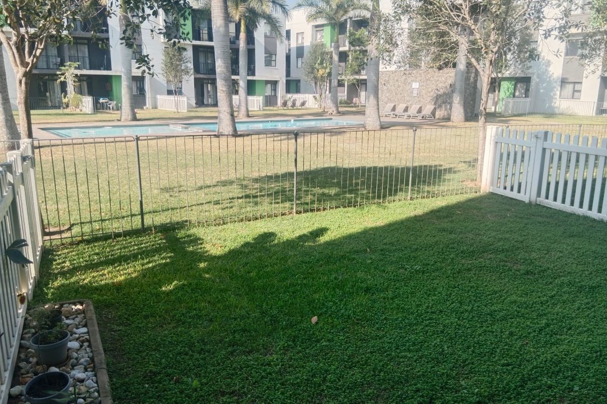 A private rear garden area featuring a well-maintained lawn, metal fencing, and direct access to the complex swimming pool and landscaped grounds.