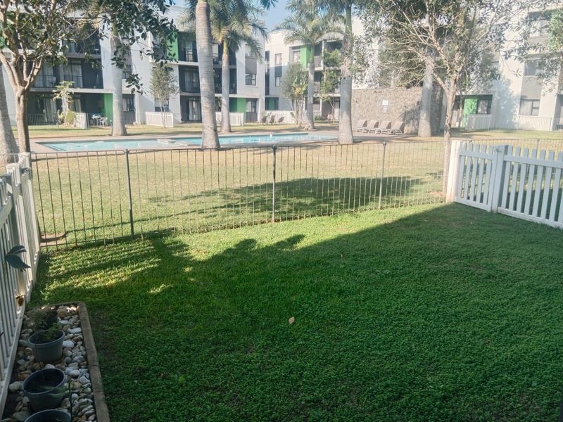 A private rear garden area featuring a well-maintained lawn, metal fencing, and direct access to the complex swimming pool and landscaped grounds.