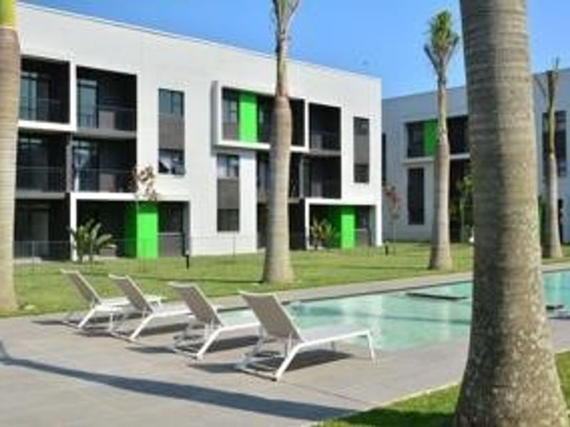 A well-maintained communal swimming pool area featuring a paved deck, landscaped garden, and modern apartment building facades with balconies.