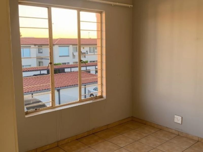 An empty bedroom featuring tiled flooring and a large window fitted with security bars, offering a view of the complex exterior.