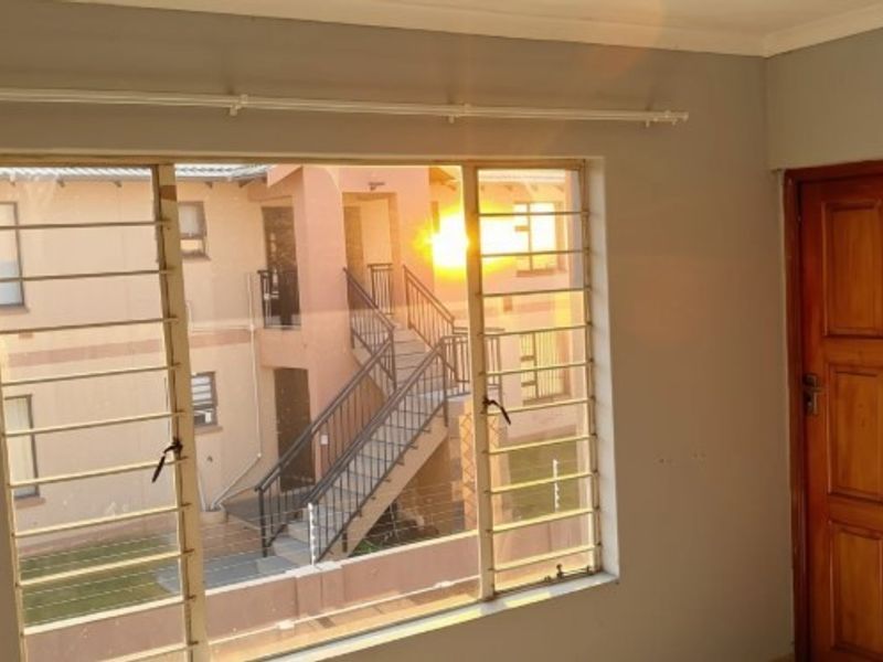 An empty bedroom featuring a timber door, curtain rail, and large windows fitted with security bars, overlooking the apartment complex courtyard.