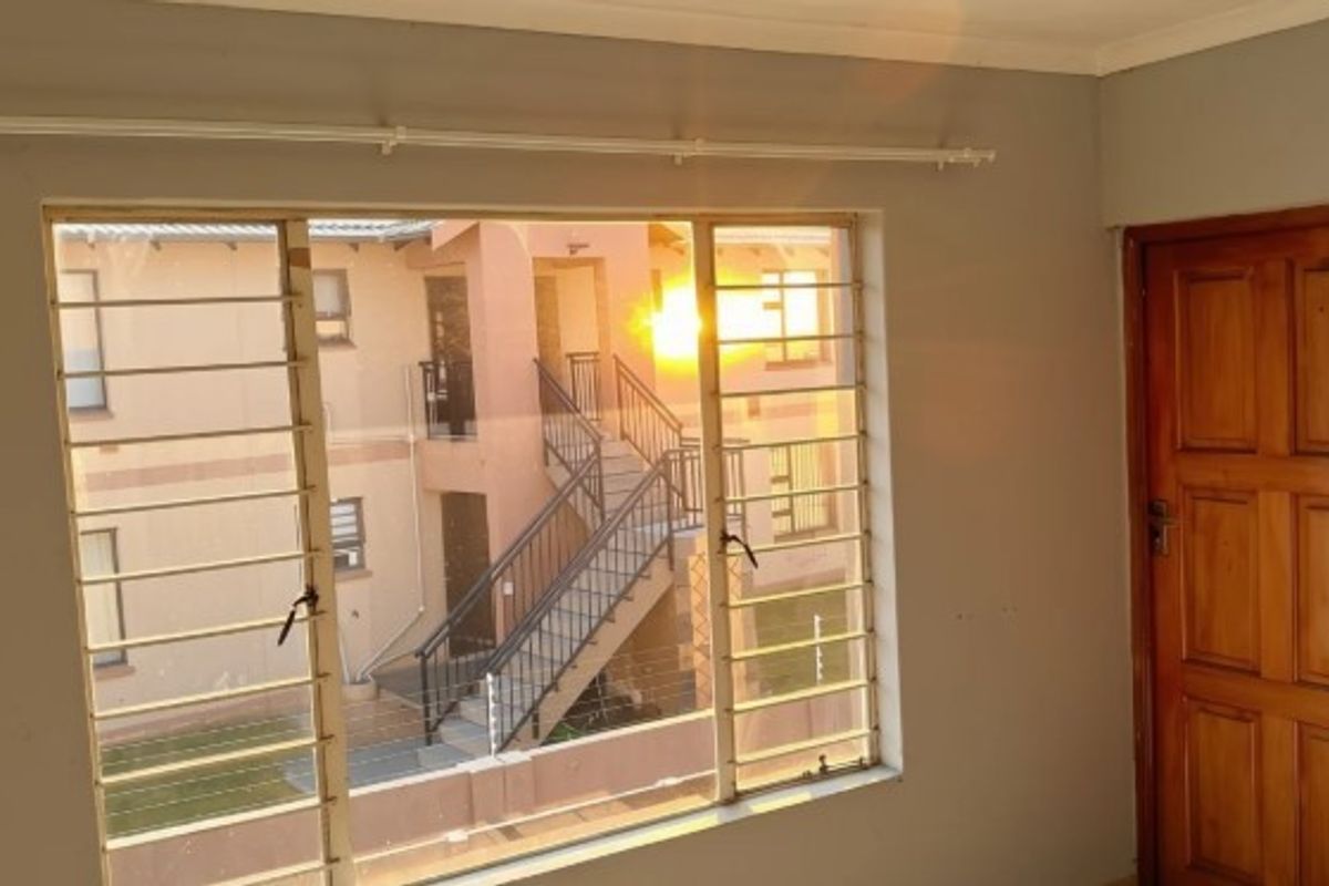An empty bedroom featuring a timber door, curtain rail, and large windows fitted with security bars, overlooking the apartment complex courtyard.