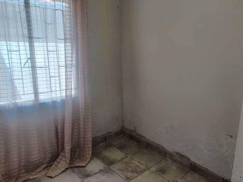 An empty bedroom featuring tiled flooring and a window with security bars, showing signs of wall dampness and floor staining requiring maintenance.