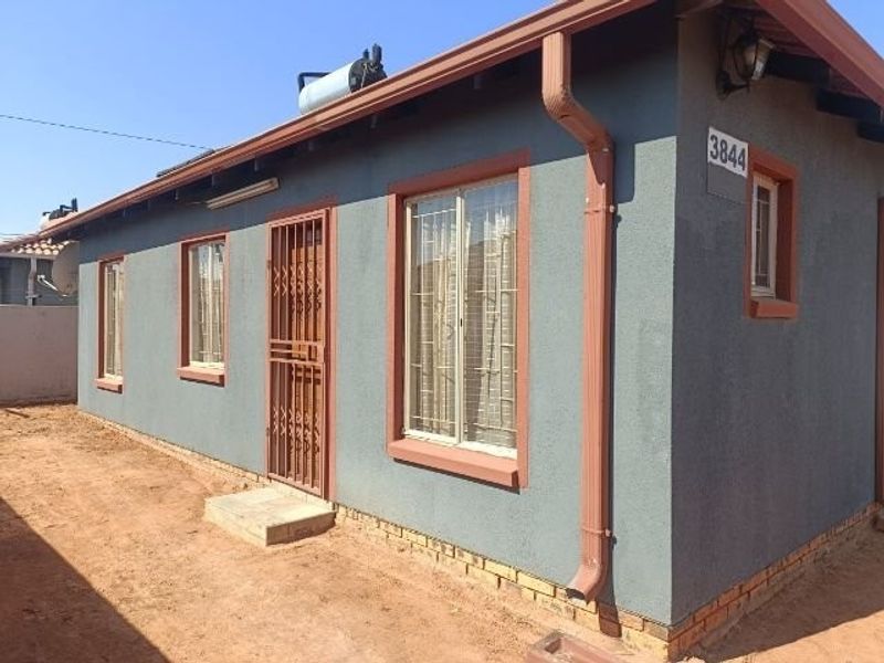 Front exterior view of a residential house featuring a solar geyser, security gates, and burglar bars on windows with a painted plaster finish.