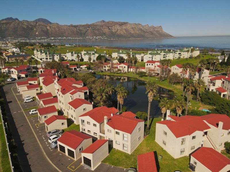 Aerial view of a residential apartment complex featuring red-tiled roofs, landscaped grounds, a central pond, and mountain views in the background.
