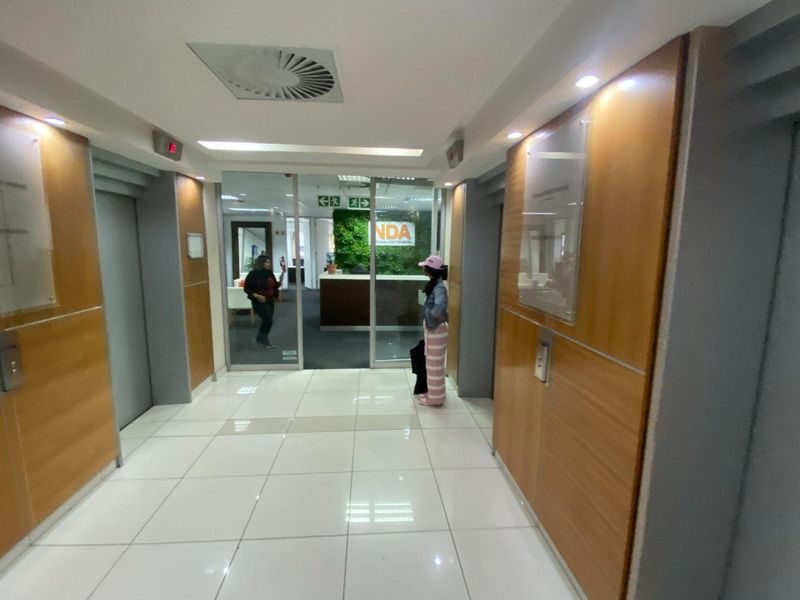 Commercial office building entrance lobby featuring elevator access, wood veneer wall paneling, and modern tiled flooring leading to a glass-enclosed reception area.
