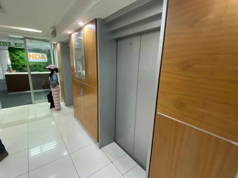 A modern commercial hallway featuring elevator access, wood veneer wall paneling, and polished tile flooring leading to a glass-partitioned office entrance.