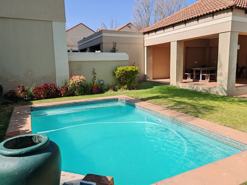 A private backyard featuring a swimming pool with tiled coping, adjacent to a covered patio area with masonry pillars and a lawn.