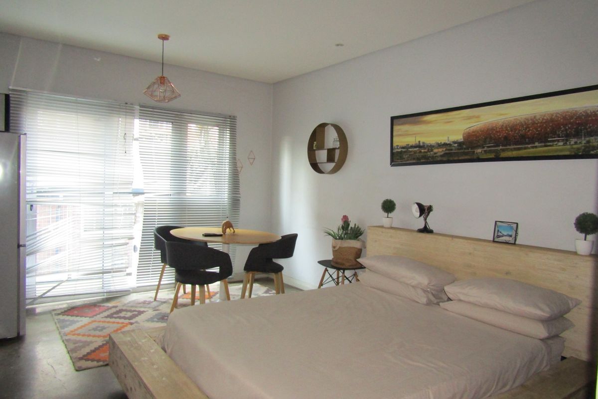 A well-lit bedroom featuring polished concrete flooring, modern pendant lighting, and large windows fitted with venetian blinds.