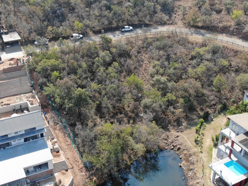 Aerial view of a sloped, undeveloped residential plot featuring natural vegetation, adjacent to existing residential structures and a small water feature.