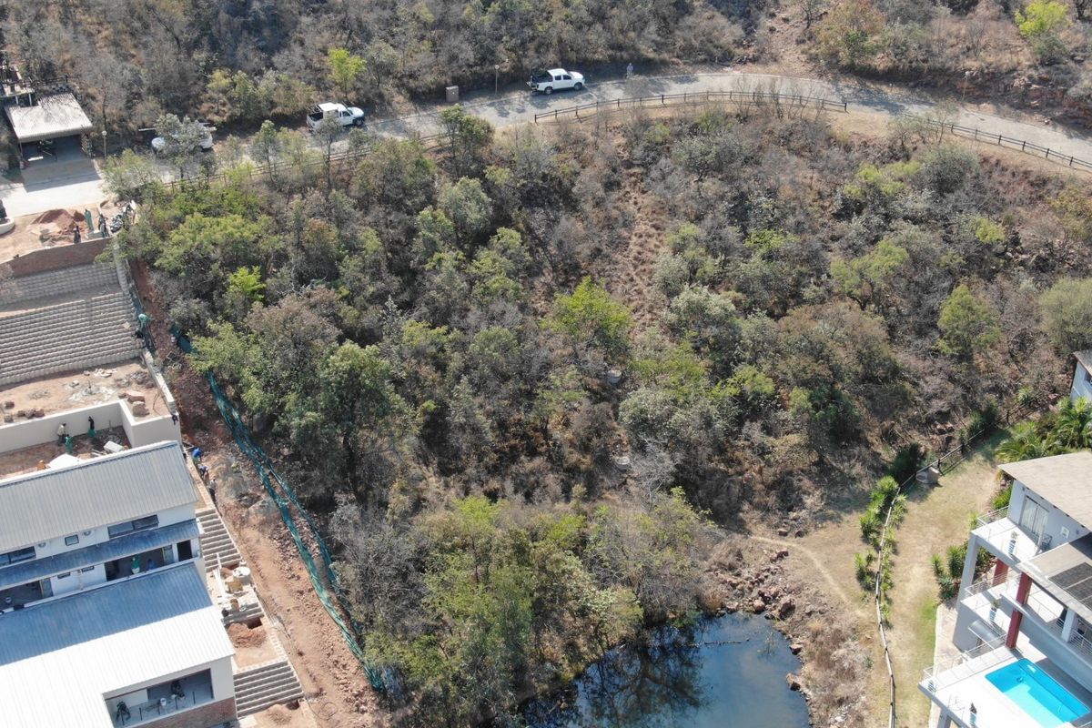 Aerial view of a sloped, undeveloped residential plot featuring natural vegetation, adjacent to existing residential structures and a small water feature.