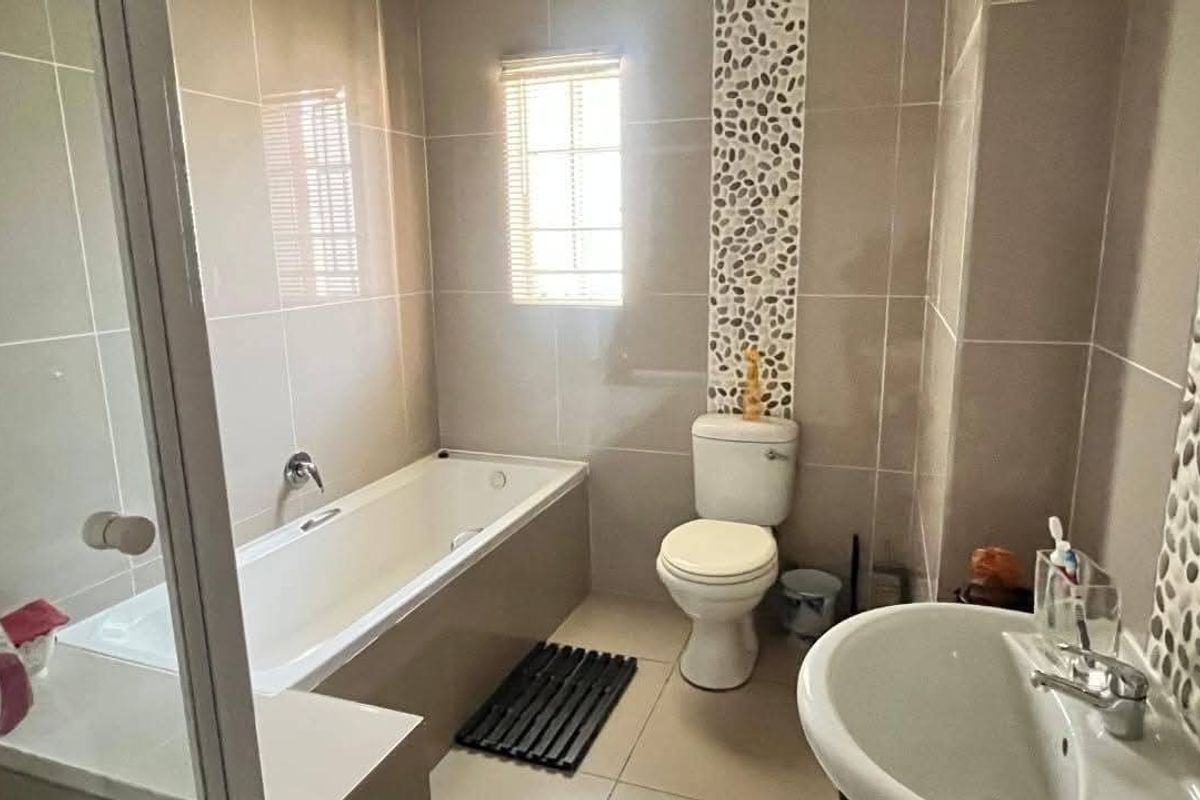 A clean, functional bathroom featuring a bathtub, pedestal basin, toilet, and decorative pebble mosaic wall accents with neutral floor-to-ceiling tiling.