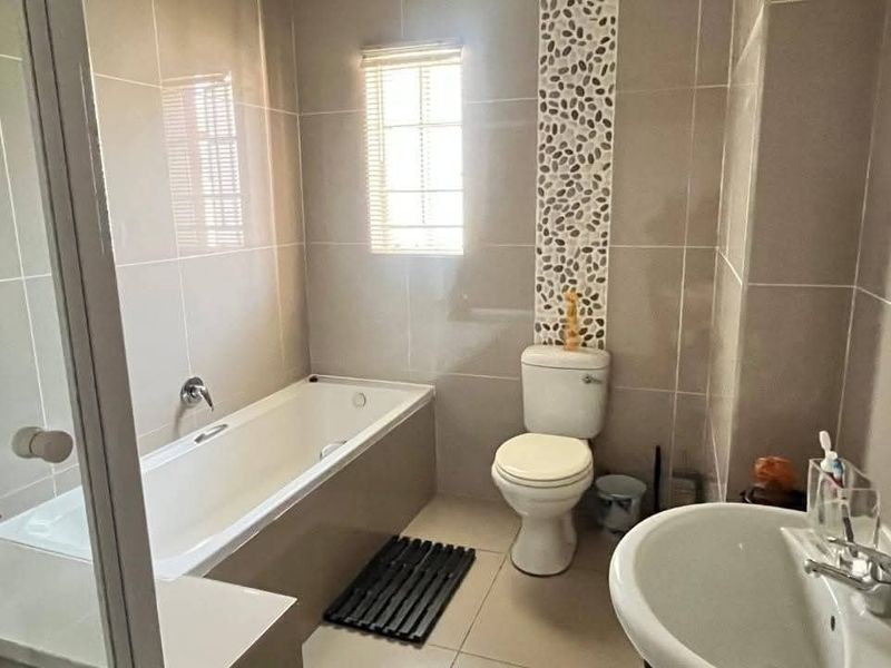 A clean, functional bathroom featuring a bathtub, pedestal basin, toilet, and decorative pebble mosaic wall accents with neutral floor-to-ceiling tiling.