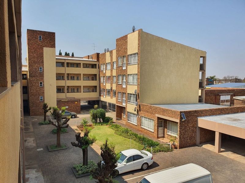 An elevated view of the apartment complex exterior, featuring a central paved courtyard, landscaped garden areas, and covered parking facilities.