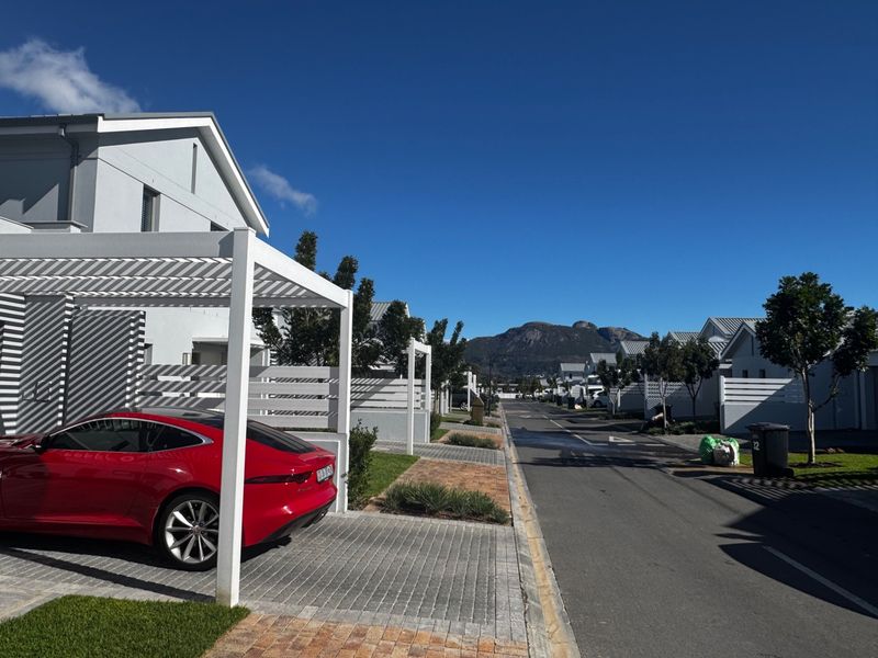 A clear street view of a modern townhouse complex featuring paved driveways, white pergolas, and well-maintained landscaping with mountain views in the background.