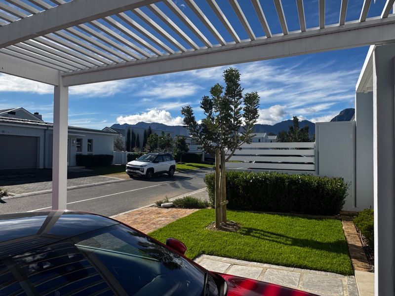 A modern outdoor area featuring a white slatted pergola, overlooking a well-maintained garden, paved driveway, and scenic mountain views.