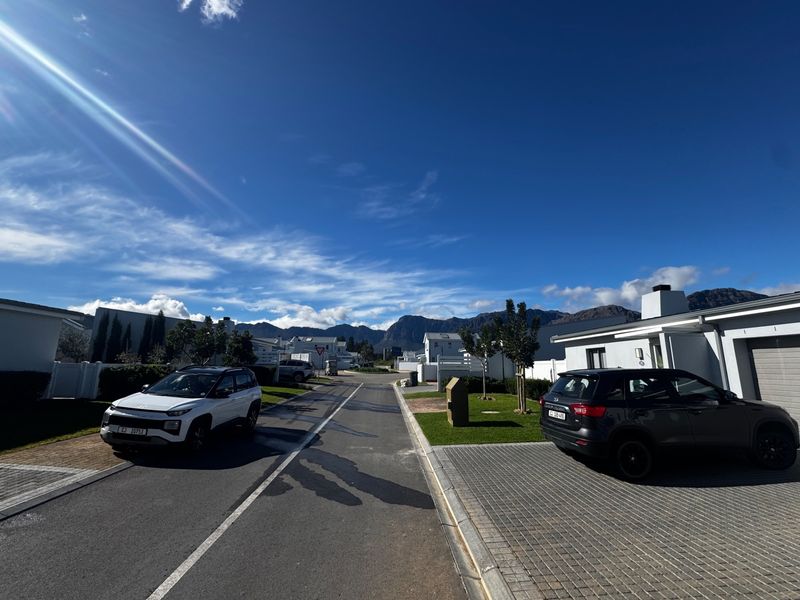 A clear street view of a modern residential townhouse estate featuring paved driveways, manicured verges, and scenic mountain views in the background.