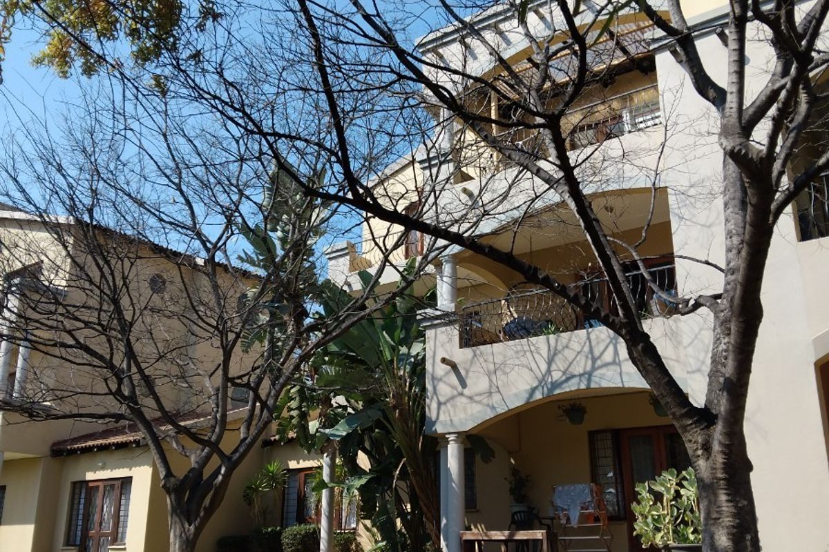 Rear view of a multi-story apartment building featuring covered balconies, ground floor patios, and established garden landscaping.
