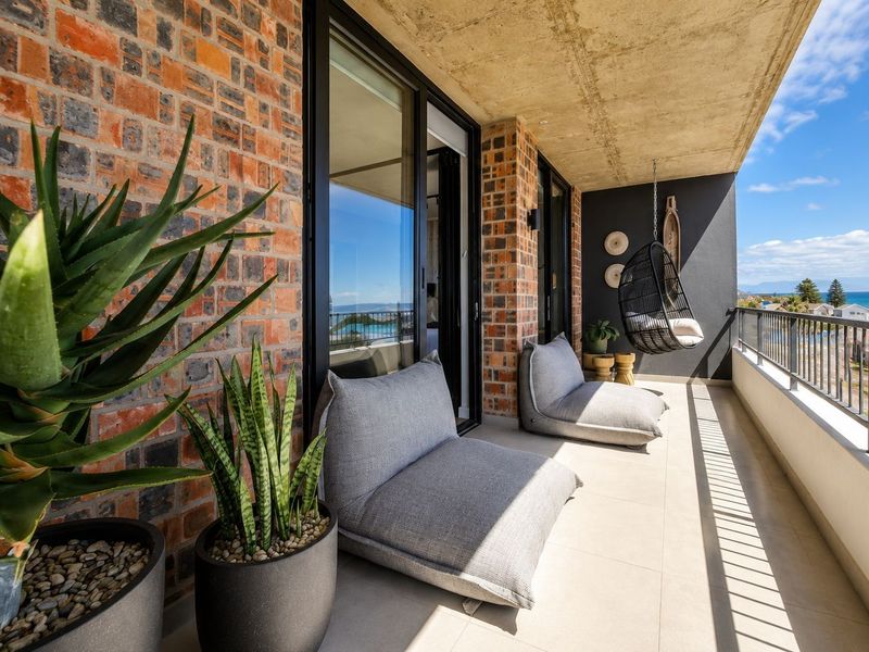 A modern covered balcony featuring exposed brickwork, concrete ceiling, and sliding glass doors, offering an outdoor space with scenic coastal views.