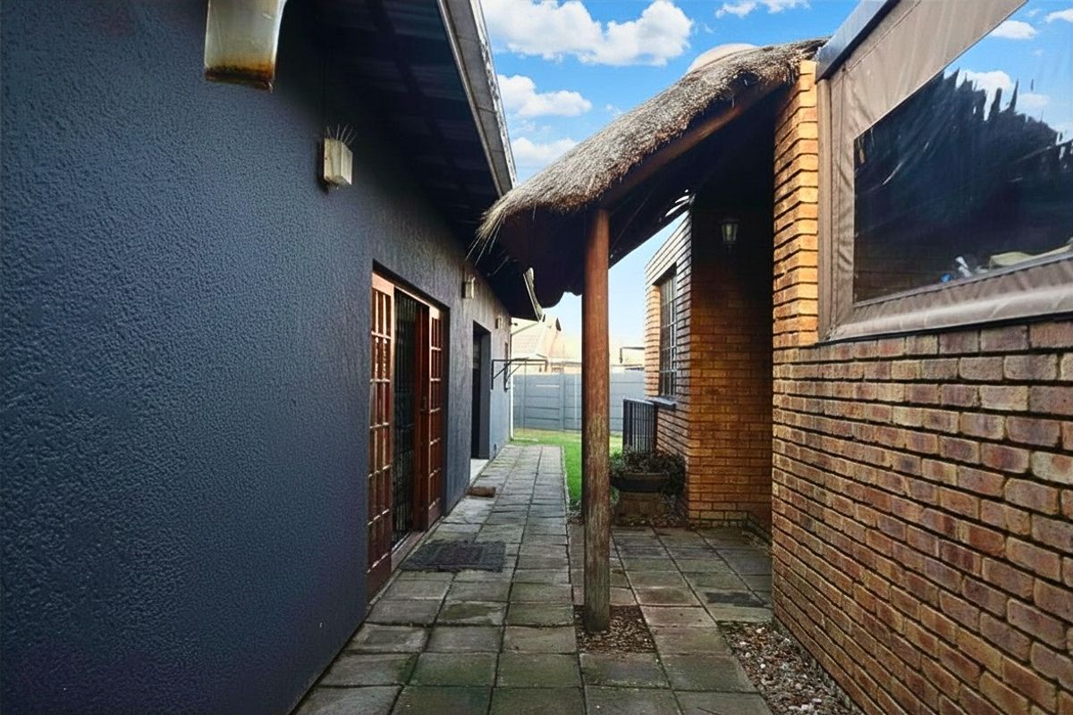 A narrow paved walkway between two building structures, featuring a thatched roof overhang and exposed brickwork.