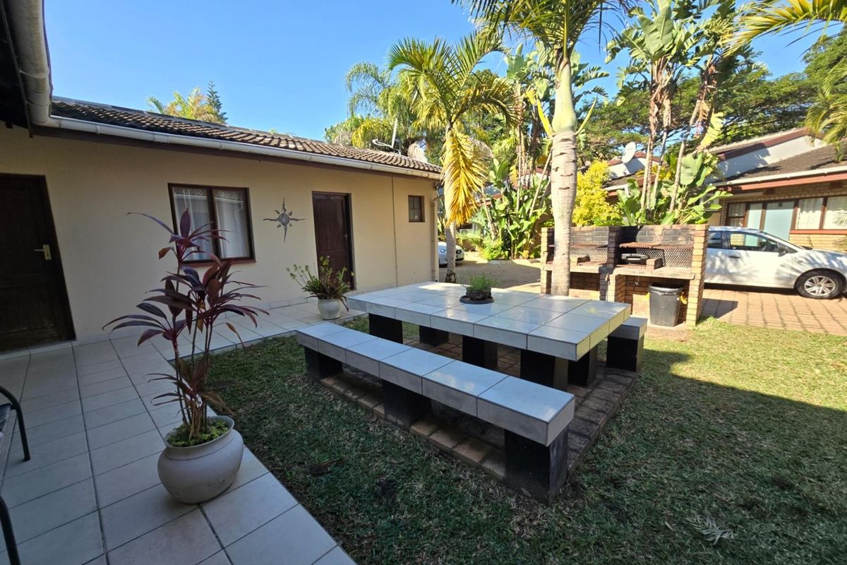Rear exterior area featuring a paved patio, built-in brick braai, and a permanent tiled outdoor dining set within a landscaped garden setting.