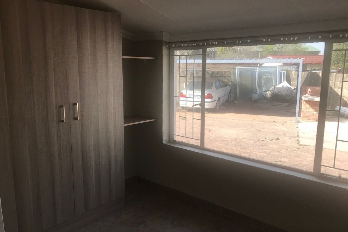 A bedroom featuring built-in wardrobes, open shelving, tiled flooring, and a large window with security bars overlooking the rear yard.